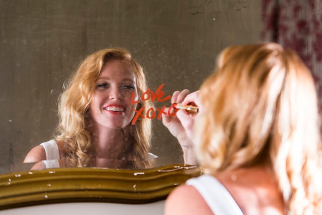 Beautiful, smiling young woman writing the word LOVE and XOXO with red lipstick on a mirror. Focus on the woman's reflection.