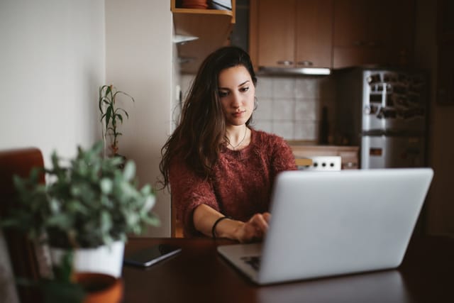 etro toned portrait of a young woman wearing casual clothing is typing on her laptop computer, sitting in the kitchen, working from her apartment space or just casually browsing the web.