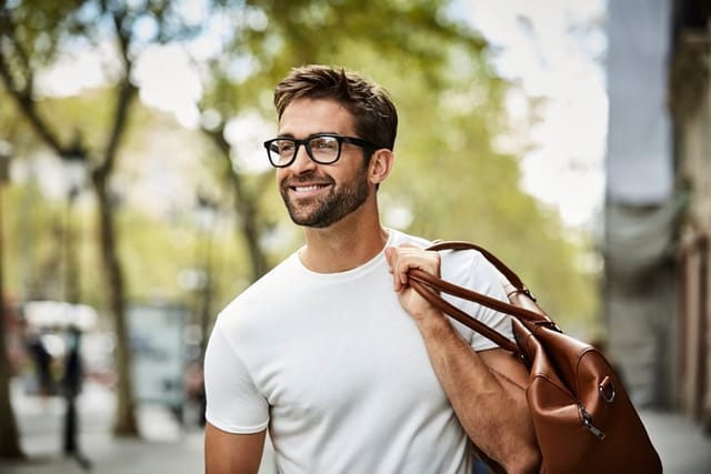 Smiling handsome businessman with beard walking in city. Executive is looking away while carrying bag. He is wearing eyeglasses.