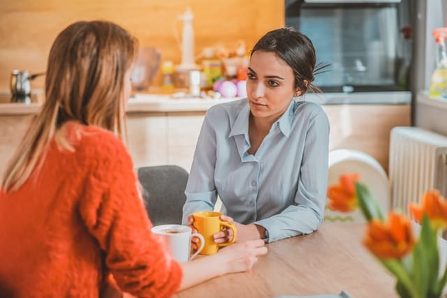 Peacefully talking. Beaming joyful women having expressive conversation during light French breakfast at home during the day. Lesbian couple lifestyle. Photo of two sad friends talking and drinking coffee and tea sitting at table in the kitchen at home. Two young beautiful Caucasian girls sitting with coffee mugs in their hands. Difficult times. Depressed cheerless woman sitting together with her partner thinking and talking about problems
