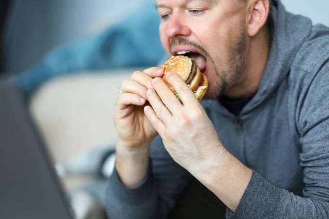 Portrait of male sitting indoors and eating big tasty hamburger full of mischievous calories and harmful cholesterol. Person consuming portion of junk food