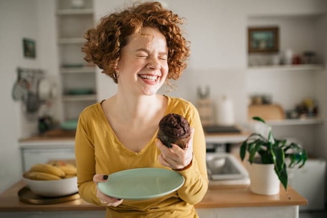 Beautiful Caucasian redhead woman with curly hair, having fun at home, smiling and relaxing, having and enjoying a cupcake desert for breakfast