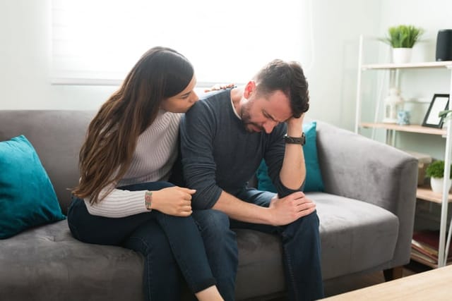 Beautiful young woman talking to her depressed boyfriend while sitting on the couch. Sad man crying and feeling upset after breaking up with his girlfriend