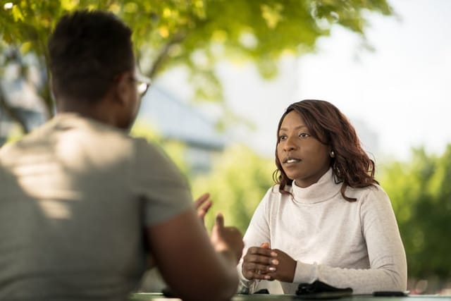 Two young adults of African decent, sit at a picnic table outside on a sunny summer day as they discuss social issues and their struggles as young Black adults. They are both dressed casually as they share their stories and concerns.