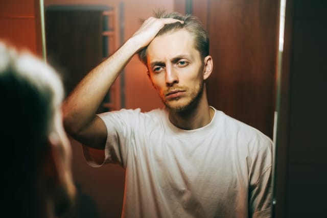 Young caucasian man standing in front mirror touching face and looking on himself.