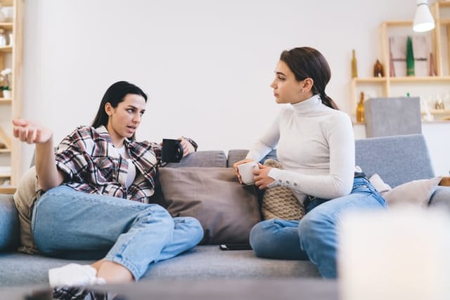 Female friends in casual wearing chatting with each other while sitting on sofa and drinking coffee in cozy living room at home