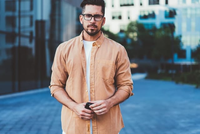 Portrait of handsome Caucasian man with modern technology for communication standing on urban setting and posing, bearded user of smartphone in optical eyewear for vision correction looking at camera