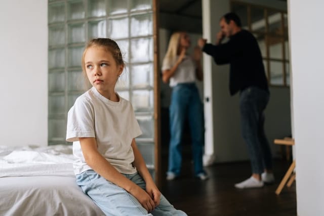 Portrait of unhappy cute little girl sadness looking away sitting on bed during parents quarrelling and fighting in living room on background. Concept of family problems, conflict, crisis.