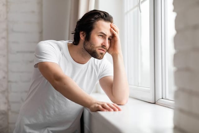 Upset young man in white t-shirt standing by window at home,