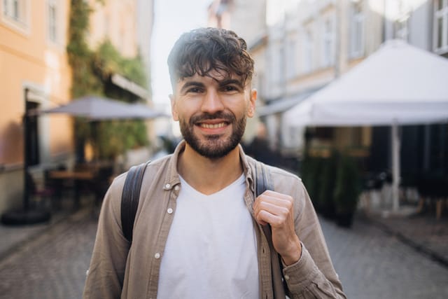 A handsome man with a warm smile stands in a charming historic European city street. His joyful expression adds a touch of charm to the picturesque surroundings.