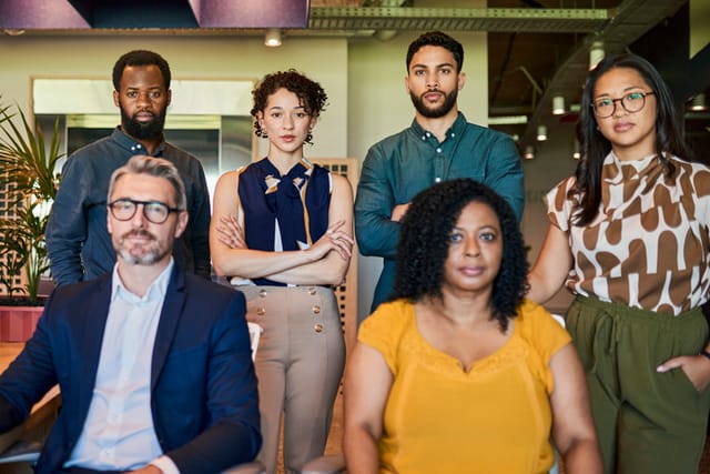 A diverse group of professionals in business attire posing together in an office setting, including a black man, black woman, white man, white woman, and woman of color. they are all smiling and looking at the camera.