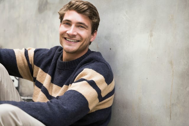 Portrait of young man smiling against wall. Handsome male is wearing sweater. He is sitting outdoors.