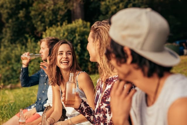 Portrait of happy young people sitting by a lake with beers. Group of friends hanging out at the lake.
