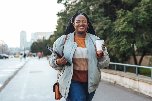 smiling black woman walking with headphones and coffee
