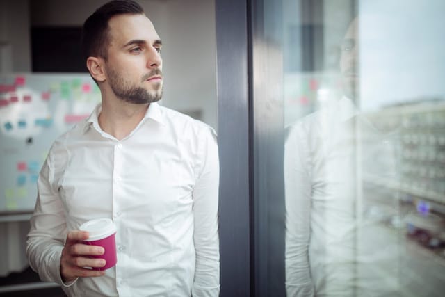 A business man in a white shirt drinking coffee and looking out the window of his office.