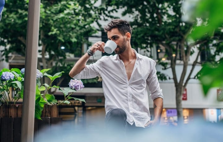 Handsome modern man with white shirt and beard, standing outdoors and drinking coffee.