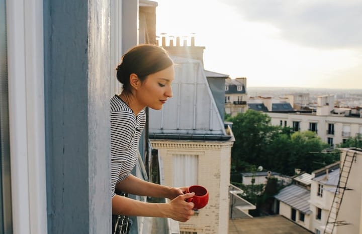 Vintage toned image of a young woman relaxing, drinking coffee, on the small balcony - window of her beautiful apartment on Montmartre, Paris. Taken in the magic hour just as the sun sets down over Parisian city scape in the background and the sunrays paint her hair bright.