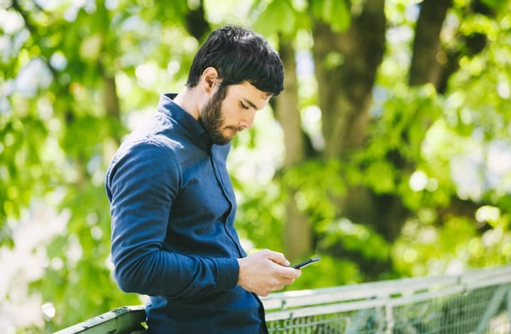 Young good-looking guy with blue shirt, dark hair and beard is reading and replying to messages and posts online on his smartphone. He looks relaxed, yet focused on what is happening online. A nice lonely afternoon spent outdoors and online. Copy space available. Made in Paris, France.
