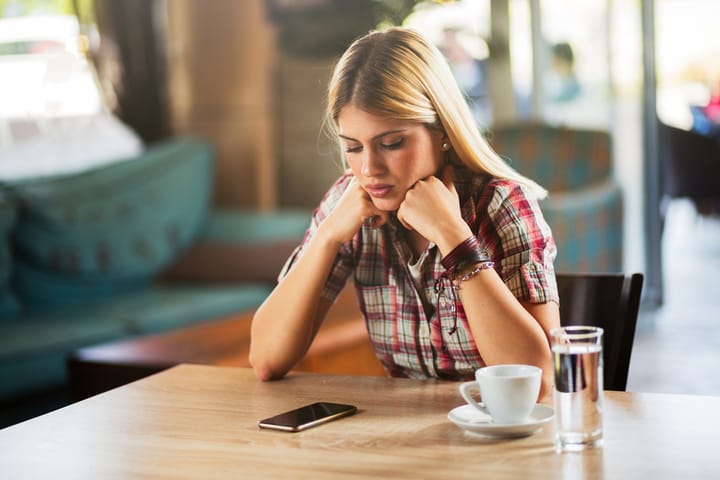 Young Pensive woman is sitting in modern cafe looking at her mobile phone waiting for a call.