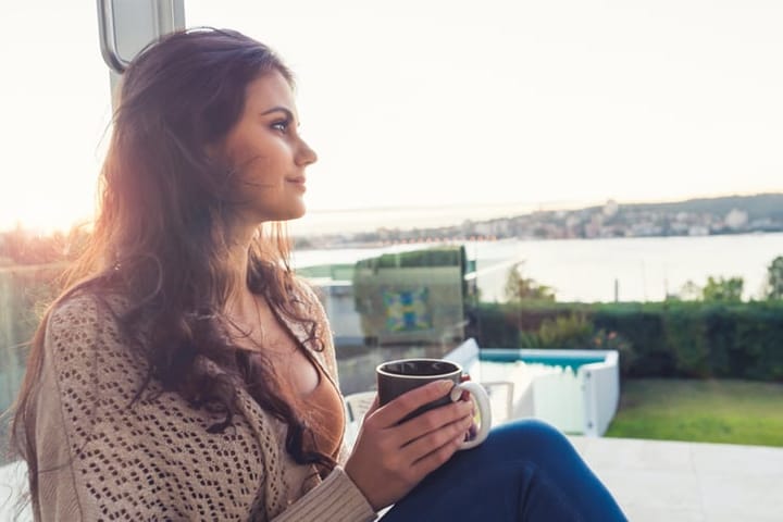 Woman drinking coffee at sunrise. She is looking at a harbour view. She is sitting on her terrace looking contemplative.