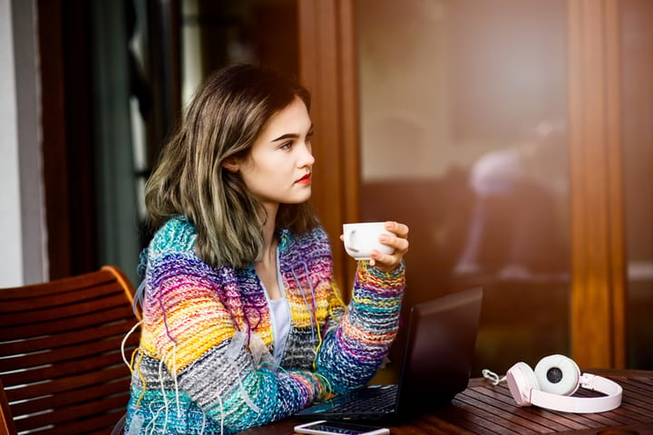 Young woman in colorful woolen sweater drinking coffee and using laptop computer on the terrace.
