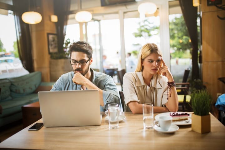 Young heterosexual couple is in a cafe on a date. Girlfriend is annoyed and displeased because her boyfriend is ignoring her and using laptop.