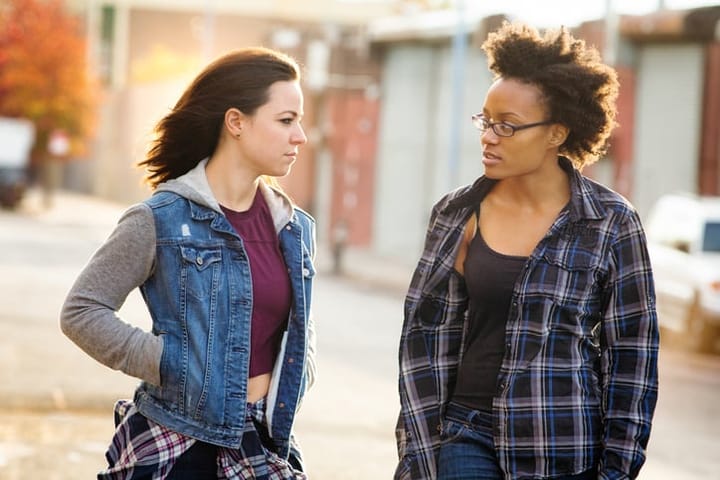 Two young female friends discuss serious matter in alley. One is black, the other white and they are both dressed in casual urban clothing. Photographed at sunset in Brooklyn.