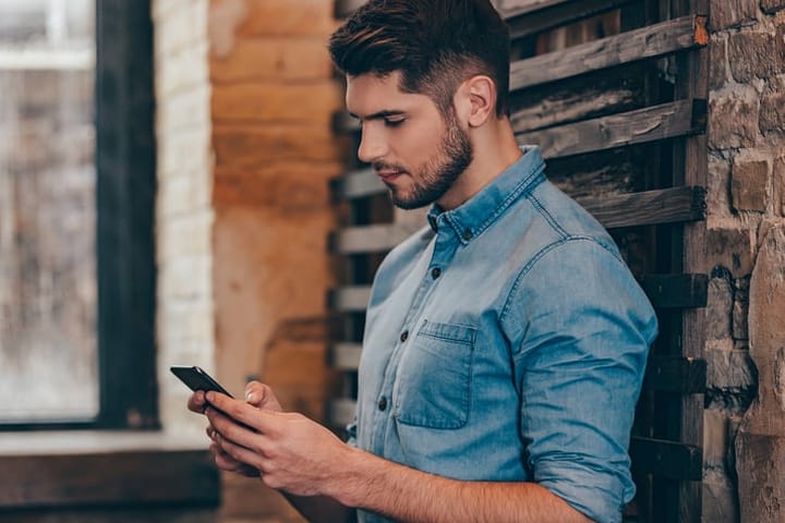 Side view of handsome thoughtful young man holding smart phone and looking at it while leaning to brick wall in loft interior