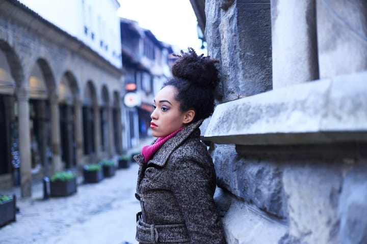 Young woman leaning on stone wall on street. Wears winter coat and pink pullover. Looking away. Stone wall with columns in a row on background. Focus on foreground.