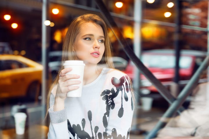 Beautiful woman drinking coffee in coffee shop from white paper coffee cup and looking to the city street through the window. Girl holding coffee cup in hand.