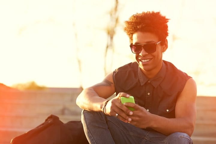 happy smiling urban hipster young man using smart phone. African american teenager holding mobile smartphone using app texting sms on sunset with lens flare