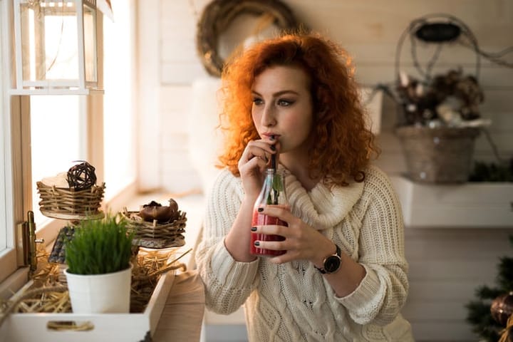 Pensive red-haired woman drinking fruit smoothie while looking away.
