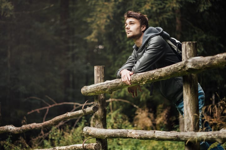 Young man with backpack hiking in the forest and leaning on a wooden fence, nature and physical exercise concept
