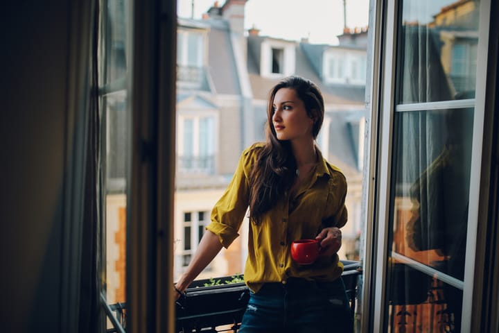 Vintage toned low key image of a young woman relaxing, drinking coffee, on the small balcony - window of her beautiful apartment on Montmartre, Paris. Taken in the magic hour just as the sun sets down, the rooftops of the Montmartre homes can be seen in the background.