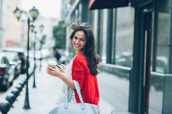 Smiling young woman with phone and coffee cup walking on the street and looking back over her shoulder.