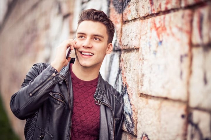Handsome young man on his phone outdoors, with a huge smile on his face, talking to someone special. Stone graffiti wall in the background.