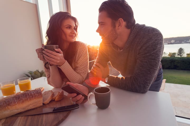 Couple having breakfast. They are drinking coffee and holding a mobile phone. They are talking. There is a water view behind them.