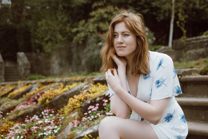 A beautiful young woman in her early 20s wearing a short white and blue dress sits in a Scottish country park on a Spring day.