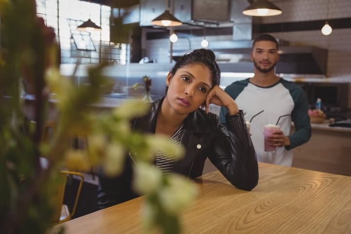 Bored woman at table with friend holding milkshake glasses in cafe