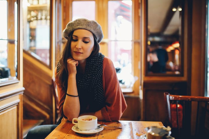 Vintage toned portrait of a young woman sitting in a traditional cafe in Paris, France, enjoying a hot cup of coffee. Shot with large aperture portrait lens for shallow depth of focus, in low, natural light conditions with added film emulation color grading. She is wearing a traditional French beret hat and a scarf, enjoying the afternoon.
