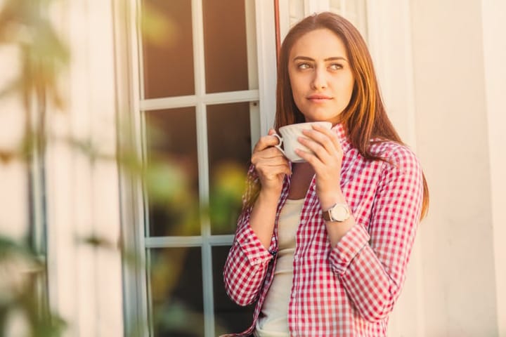 Beautiful young woman holding a cup of coffee looking through a house window.