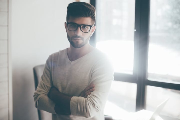Confident young man in eyeglasses keeping arms crossed and looking at camera while leaning at the desk in office