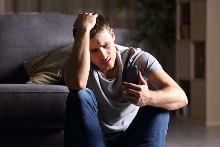 Single sad man checking mobile phone sitting on the floor in the living room at home with a dark background
