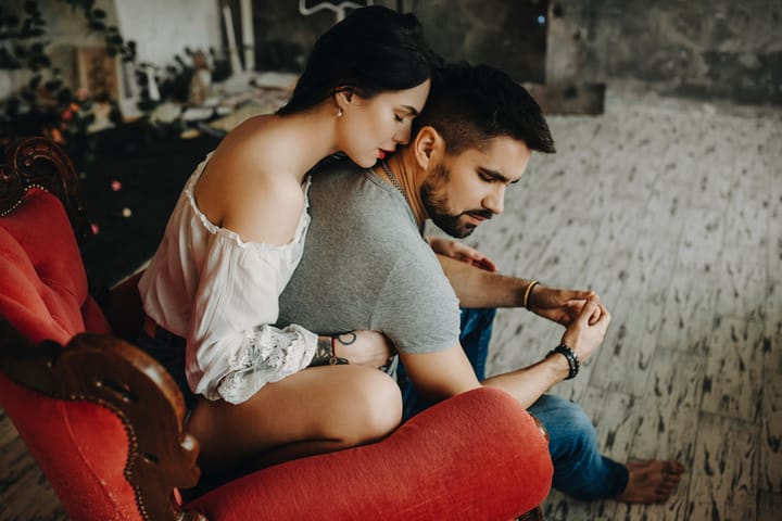 Very beautiful couple sitting on vintage red chair, where tattooed with red lips girl is embracing from back his lover waiting to kiss his neck while he is turning his head to her gently.