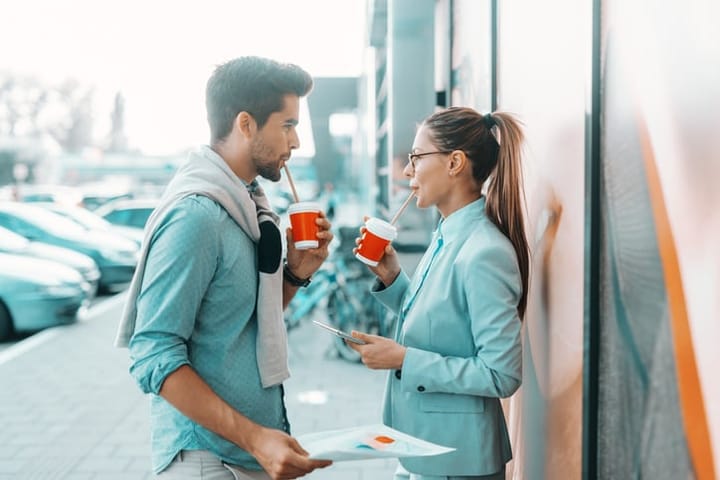 Positive young businesspeople standing outdoors, discussing and drinking coffee to go. Woman holding tablet and man holding paperwork.