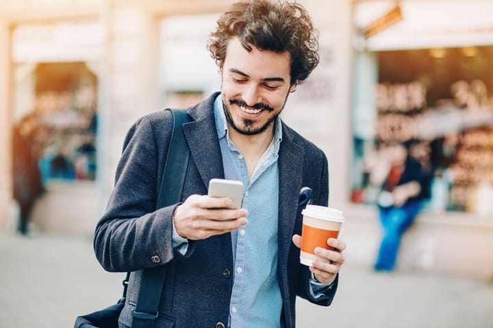 Smiling young man holding a coffee cup text messaging outside in city environment.