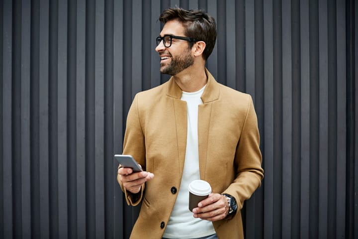 Smiling businessman with smart phone and disposable cup. Handsome executive looking away while standing against wall. He is wearing smart casuals.