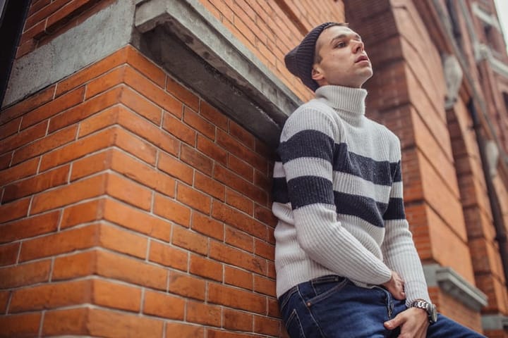 Young man in turtleneck sweater and knit hat standing on the street, feeling sad.