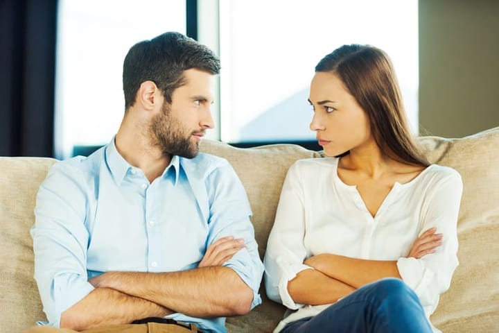 Angry young couple looking at each other and keeping arms crossed while sitting close to each other on the couch