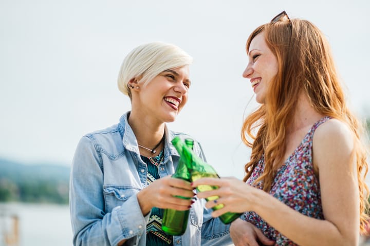 Two girlfriends spending sunny summer day at the beach, drinking refreshing cider or beer and having fun together.